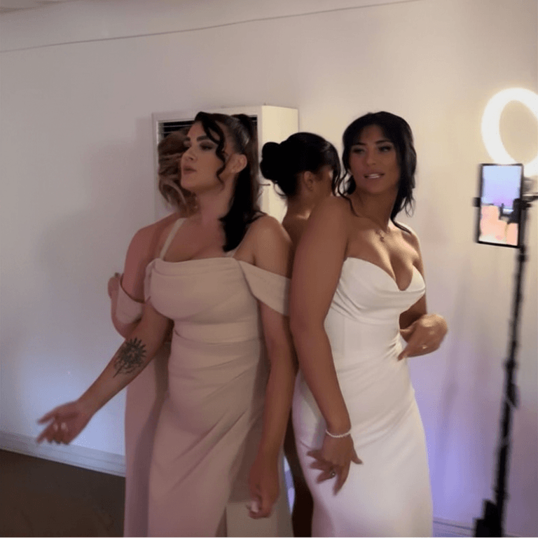 Three women in elegant dresses posing at a Smiles Better 360 video photobooth at a wedding